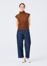 A person stands smiling against a plain background, wearing me ISSEY MIYAKE's STRETCH PLEATS 4 blue pants with stretch pleats, a sleeveless brown high-neck top, and white slip-on shoes, hands relaxed behind their back.