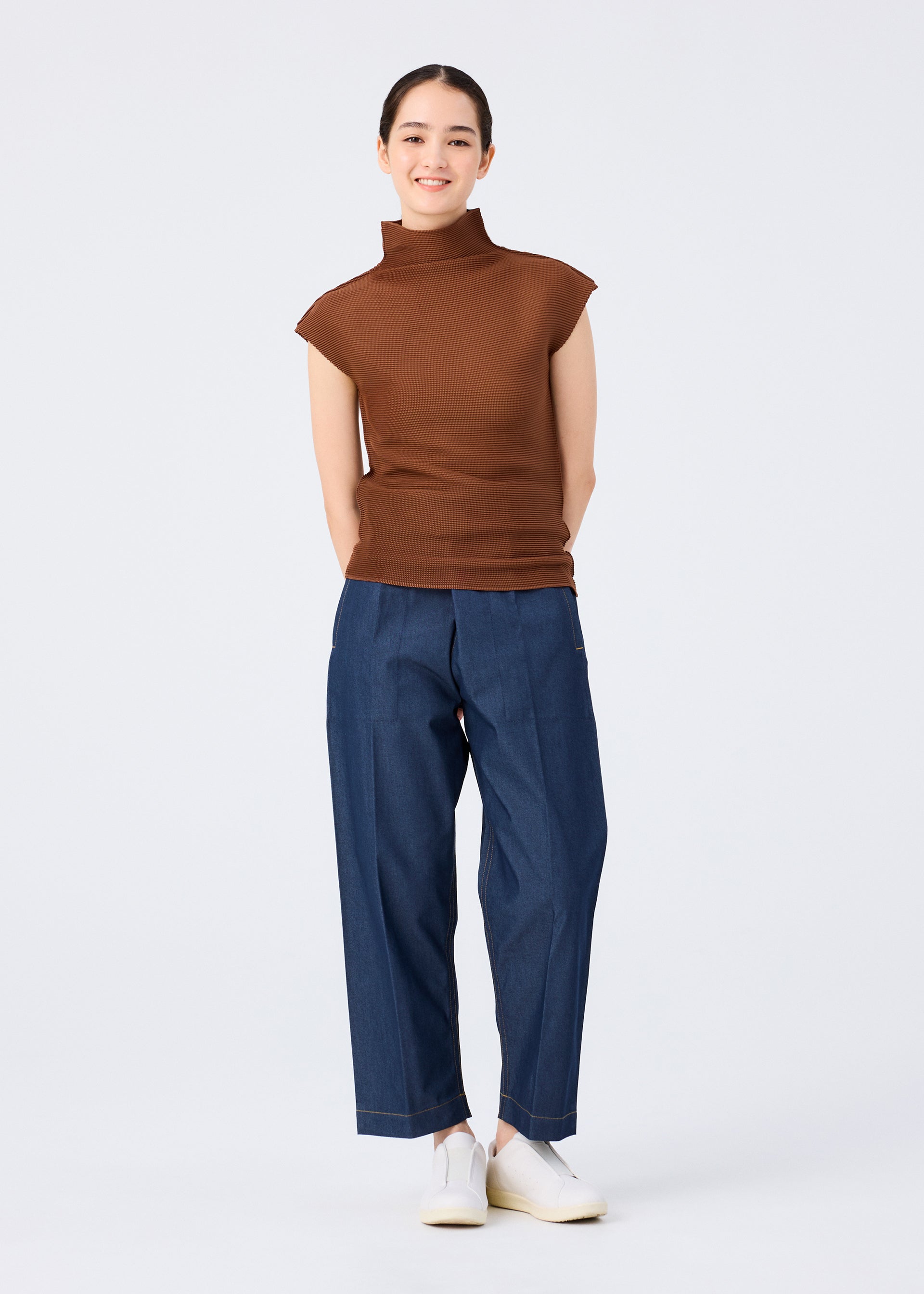 Zoomed view A person stands smiling against a plain background, wearing me ISSEY MIYAKE's STRETCH PLEATS 4 blue pants with stretch pleats, a sleeveless brown high-neck top, and white slip-on shoes, hands relaxed behind their back.
