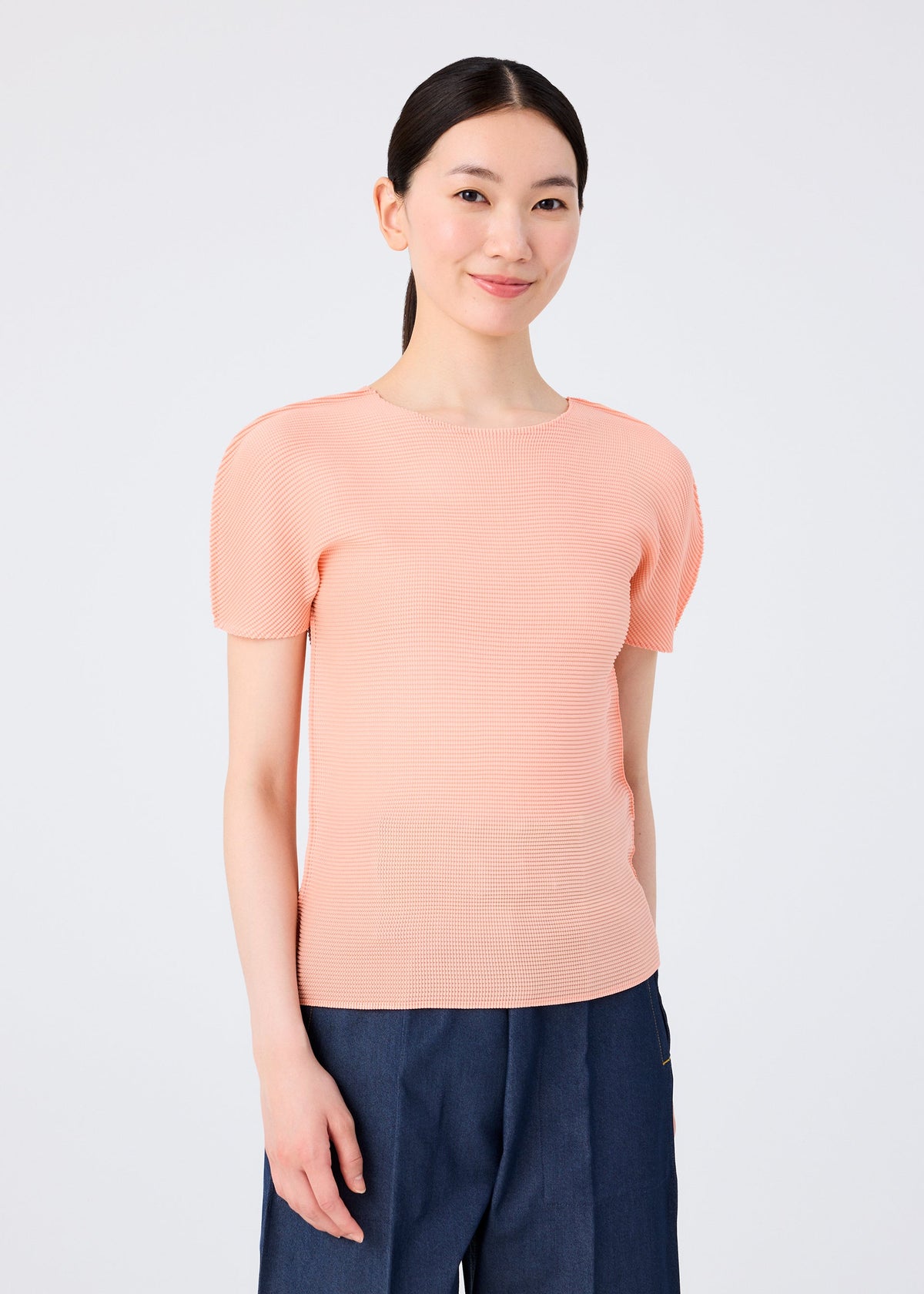 A person with long dark hair smiles against a plain white background, wearing the me ISSEY MIYAKE STRETCH PLEATS 4 light peach round-neck short-sleeved top and dark blue pants. view 5 of 7