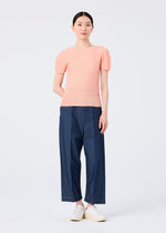 A woman stands against a white background wearing the me ISSEY MIYAKE STRETCH PLEATS 4 peach round-neck, short-sleeved top, loose blue denim pants, and white slip-on shoes. Her straight dark hair is tied back and she has a neutral expression.