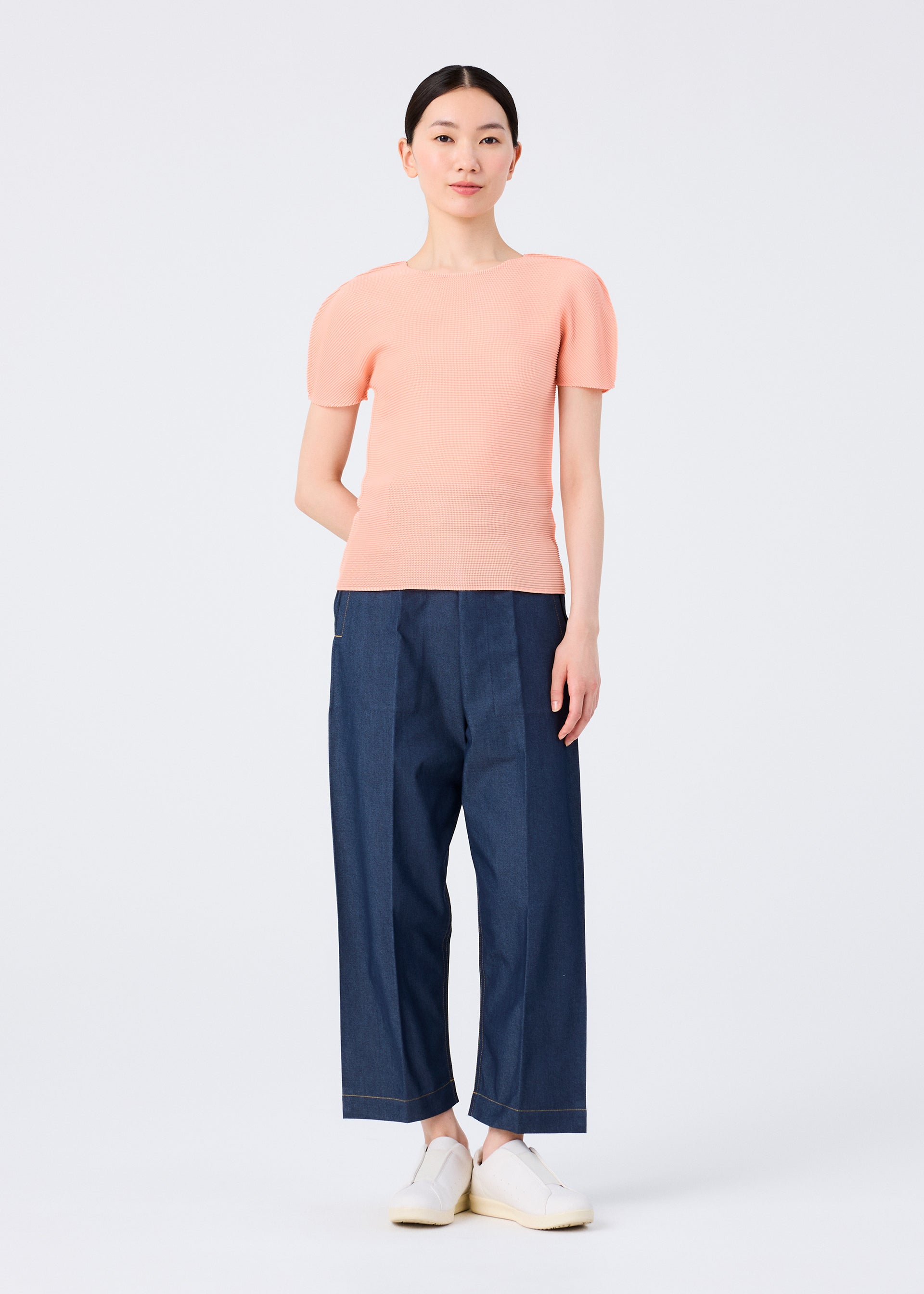 Zoomed view A woman stands against a white background wearing the me ISSEY MIYAKE STRETCH PLEATS 4 peach round-neck, short-sleeved top, loose blue denim pants, and white slip-on shoes. Her straight dark hair is tied back and she has a neutral expression.