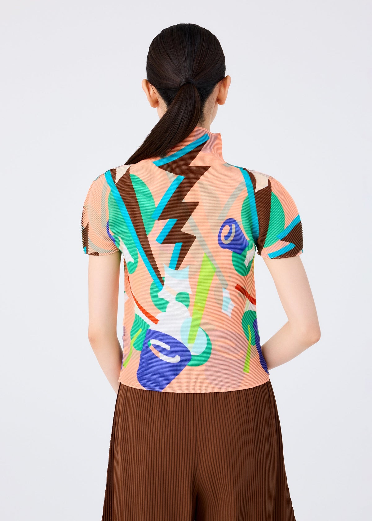 A woman with long dark hair tied back is shown from behind against a plain light background, wearing the SURPRISE BOX short-sleeved, high-neck top by me ISSEY MIYAKE with a colorful jack-in-the-box pattern and a pleated brown skirt. view 6 of 7
