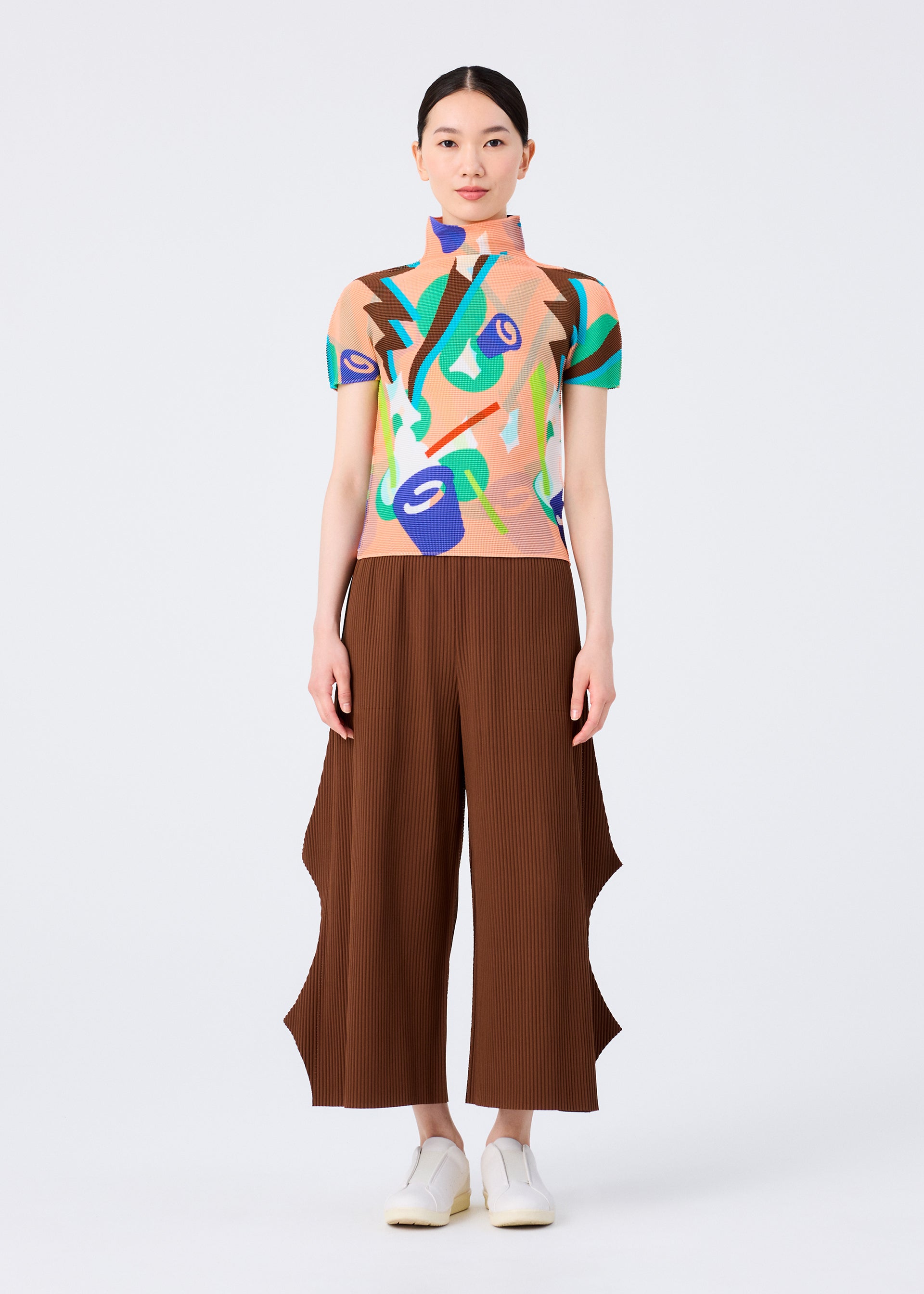 Zoomed view A woman stands facing forward against a plain white background, wearing the me ISSEY MIYAKE SURPRISE BOX high-neck top with a colorful abstract jack-in-the-box pattern, brown wide-leg pants with wavy sides, and white shoes.