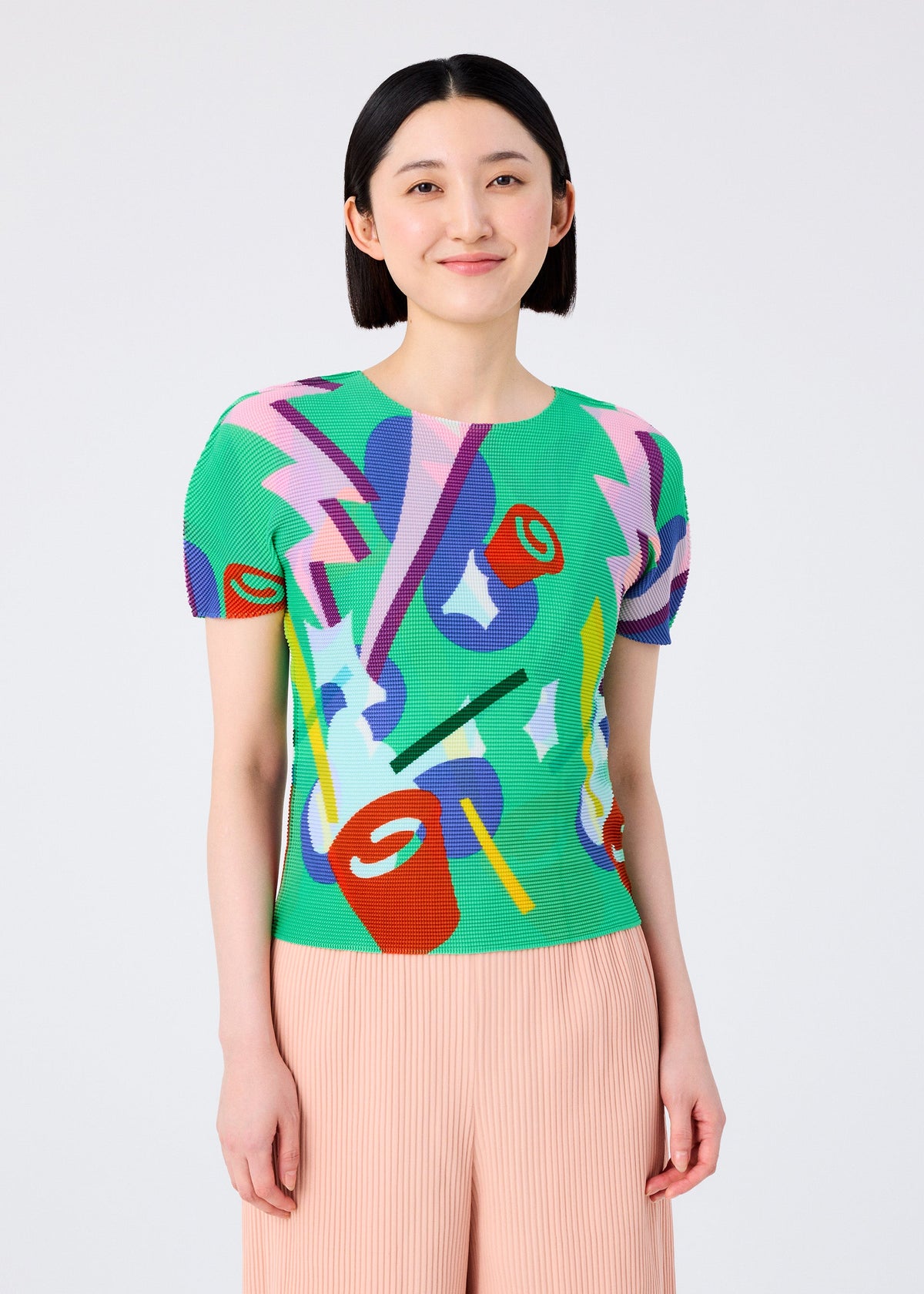 A smiling woman with short black hair wears the me ISSEY MIYAKE SURPRISE BOX—a bright, round-neck short-sleeve top with abstract shapes and lightning bolts—paired with light pink pleated pants, standing against a plain background. view 5 of 8
