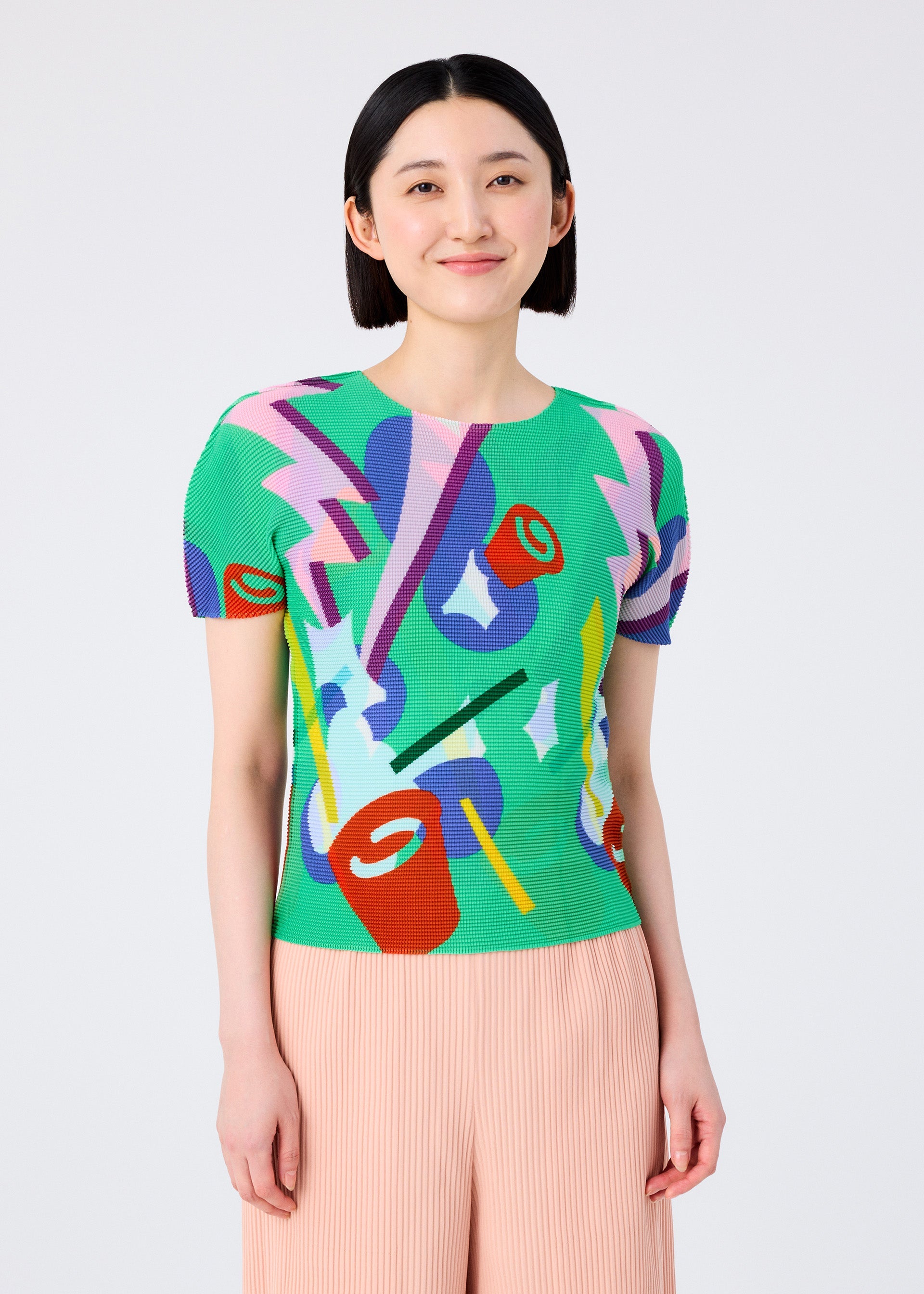 Zoomed view A smiling woman with short black hair wears the me ISSEY MIYAKE SURPRISE BOX—a bright, round-neck short-sleeve top with abstract shapes and lightning bolts—paired with light pink pleated pants, standing against a plain background.