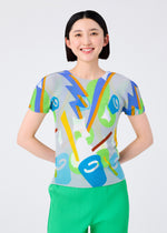 A smiling woman with short dark hair models the me ISSEY MIYAKE SURPRISE BOX: a round-neck, short-sleeved top with a colorful abstract pattern and bright green pleated pants, standing against a plain light background.