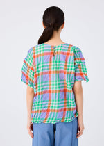 A woman with dark hair in a low ponytail is seen from behind, wearing the me ISSEY MIYAKE CHECK WHIPPED PLEATS SHIRT and light blue pants, set against a plain white background.