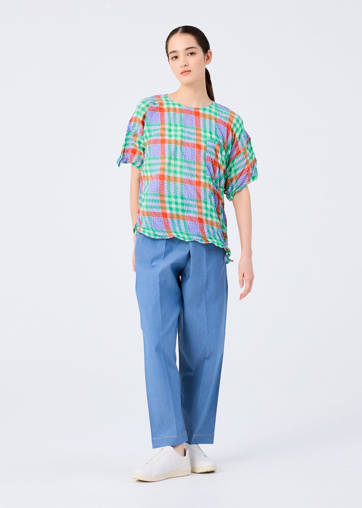 A person with long dark hair pulled back and a neutral expression wears the me ISSEY MIYAKE CHECK WHIPPED PLEATS SHIRT against a plain background, paired with light blue wide-leg pants and white slip-on shoes. view 7 of 7