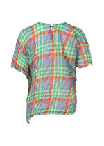 The CHECK WHIPPED PLEATS SHIRT by me ISSEY MIYAKE is a loose-fit, short-sleeved blouse made from yarn-dyed checkered fabric in green, orange, blue, and white plaid, featuring a crinkled texture and an uneven back hem.