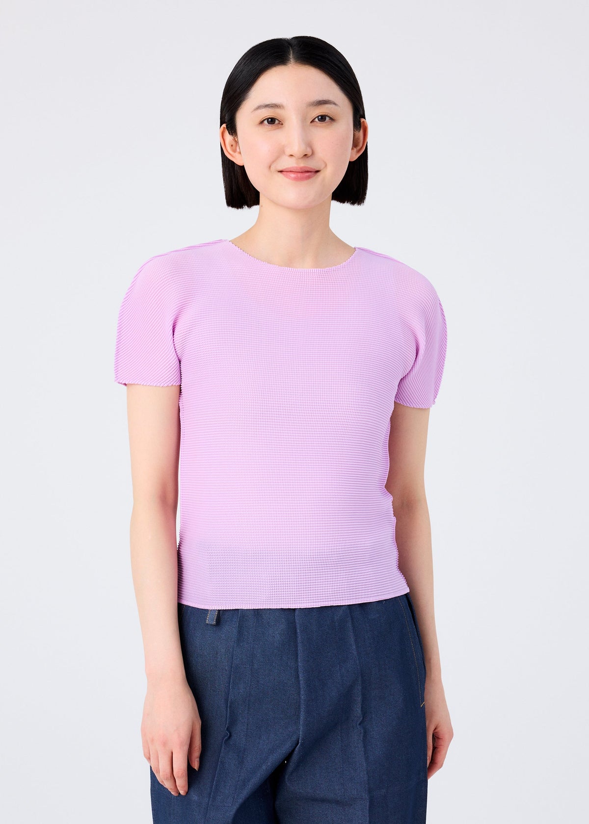 A person with straight black hair, wearing the me ISSEY MIYAKE STRETCH PLEATS 5 light pink short-sleeved top and dark blue pants, stands facing the camera against a plain white background, smiling gently. view 3 of 5