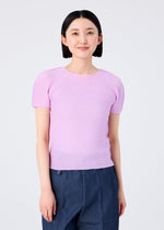 A person with straight black hair, wearing the me ISSEY MIYAKE STRETCH PLEATS 5 light pink short-sleeved top and dark blue pants, stands facing the camera against a plain white background, smiling gently.