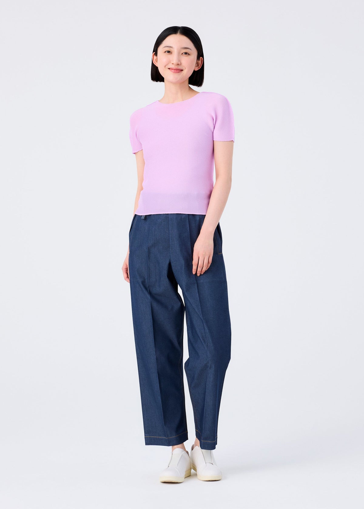 A woman smiles against a plain white background, wearing the me ISSEY MIYAKE STRETCH PLEATS 5 light purple top paired with loose-fitting blue jeans and white shoes. view 5 of 5