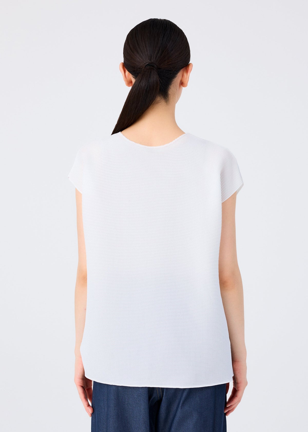 A person with long dark hair in a low ponytail is seen from behind, wearing the me ISSEY MIYAKE SQUARE STRETCH PLEATS top and blue pants against a plain white background. view 6 of 7