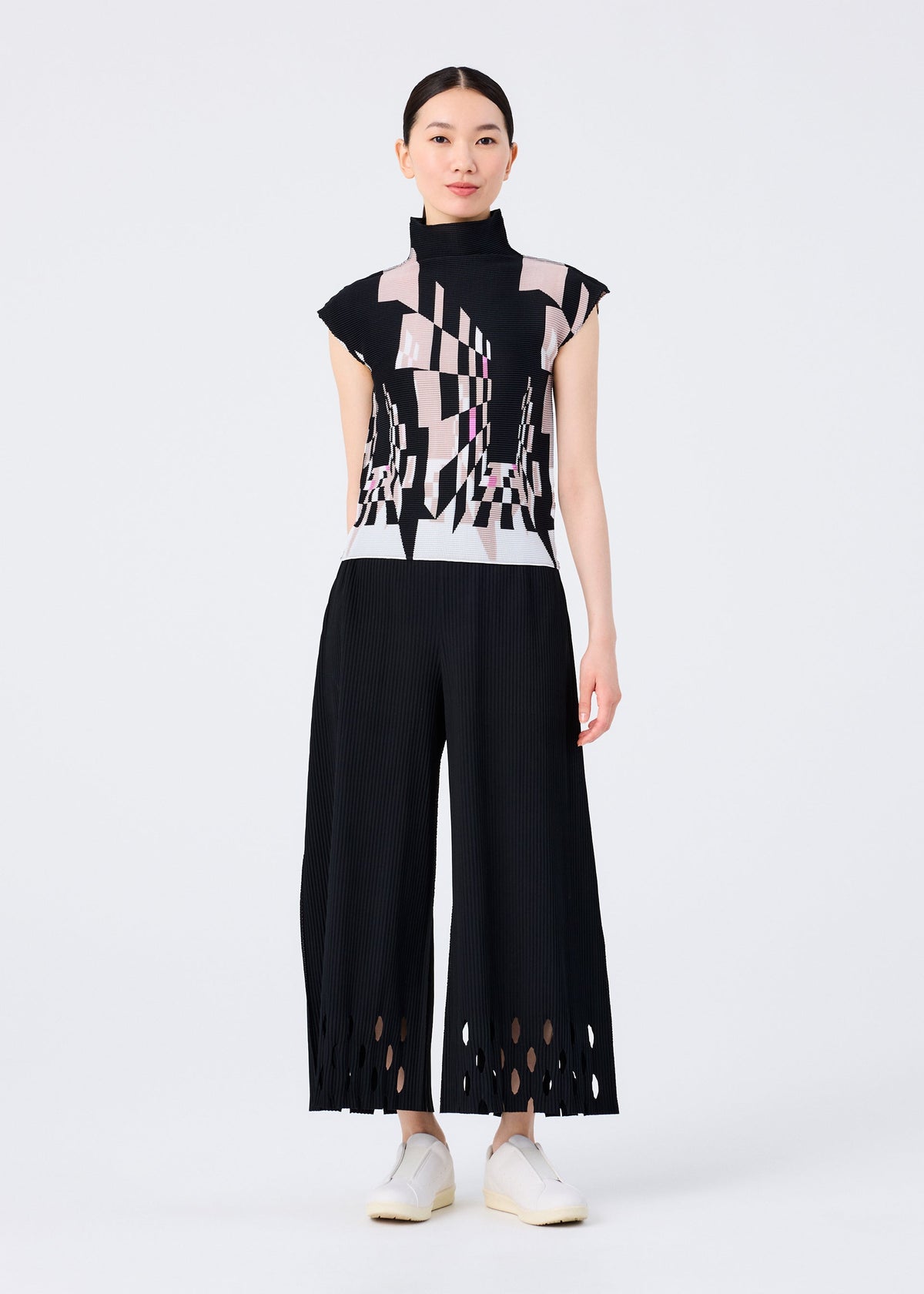 A woman faces forward wearing the me ISSEY MIYAKE PERSPECTIVE high-neck top with a black, pink, and white geometric pattern and French sleeves, paired with black STRETCH PLEATS wide-leg pants featuring cut-out hems. view 6 of 6