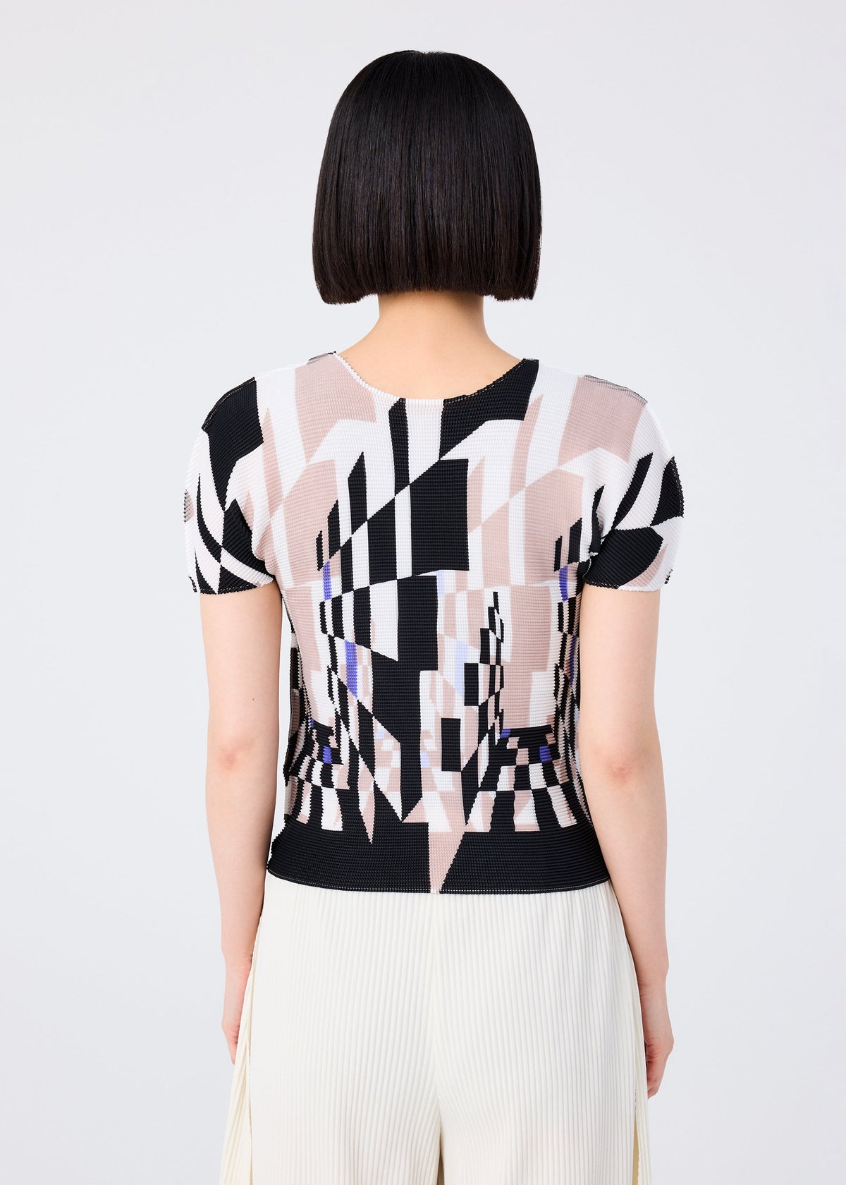 A person with short, straight black hair is seen from behind wearing the me ISSEY MIYAKE PERSPECTIVE patterned top in black, beige, and white with a round neck and short sleeves, paired with cream STRETCH PLEATS pants against a plain background. view 5 of 6