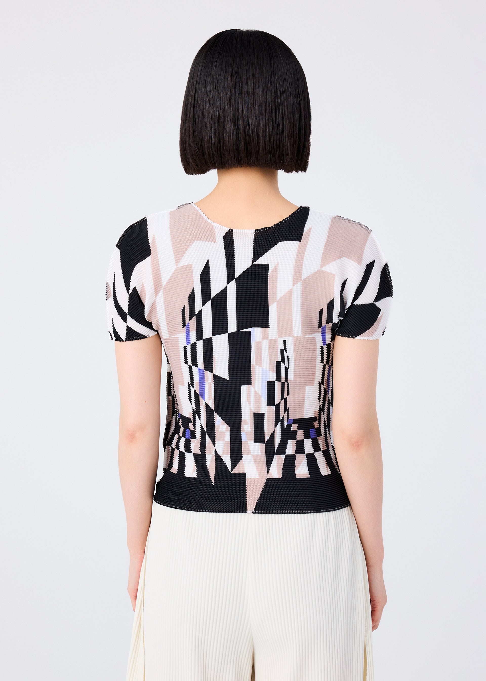 Zoomed view A person with short, straight black hair is seen from behind wearing the me ISSEY MIYAKE PERSPECTIVE patterned top in black, beige, and white with a round neck and short sleeves, paired with cream STRETCH PLEATS pants against a plain background.
