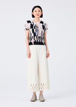 A woman faces forward against a white background, smiling in the me ISSEY MIYAKE PERSPECTIVE round-neck pleated top with geometric patterns and wide-leg cream pants featuring hem cut-outs, paired with beige shoes.