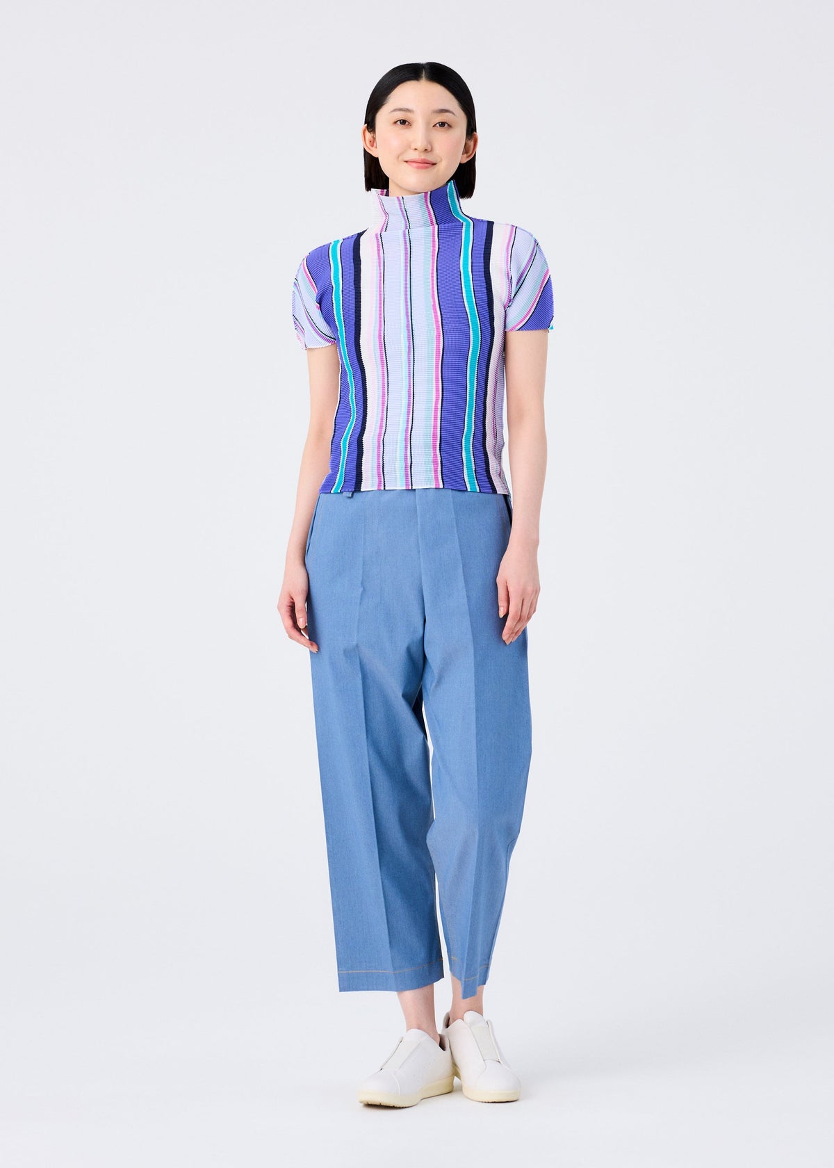 A person stands smiling in a me ISSEY MIYAKE SHIRT STRIPE high-neck, short-sleeved top with blue, green, and purple stripes, paired with loose light blue pants and white shoes against a plain white background. view 6 of 6