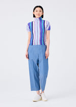 A person stands smiling in a me ISSEY MIYAKE SHIRT STRIPE high-neck, short-sleeved top with blue, green, and purple stripes, paired with loose light blue pants and white shoes against a plain white background.