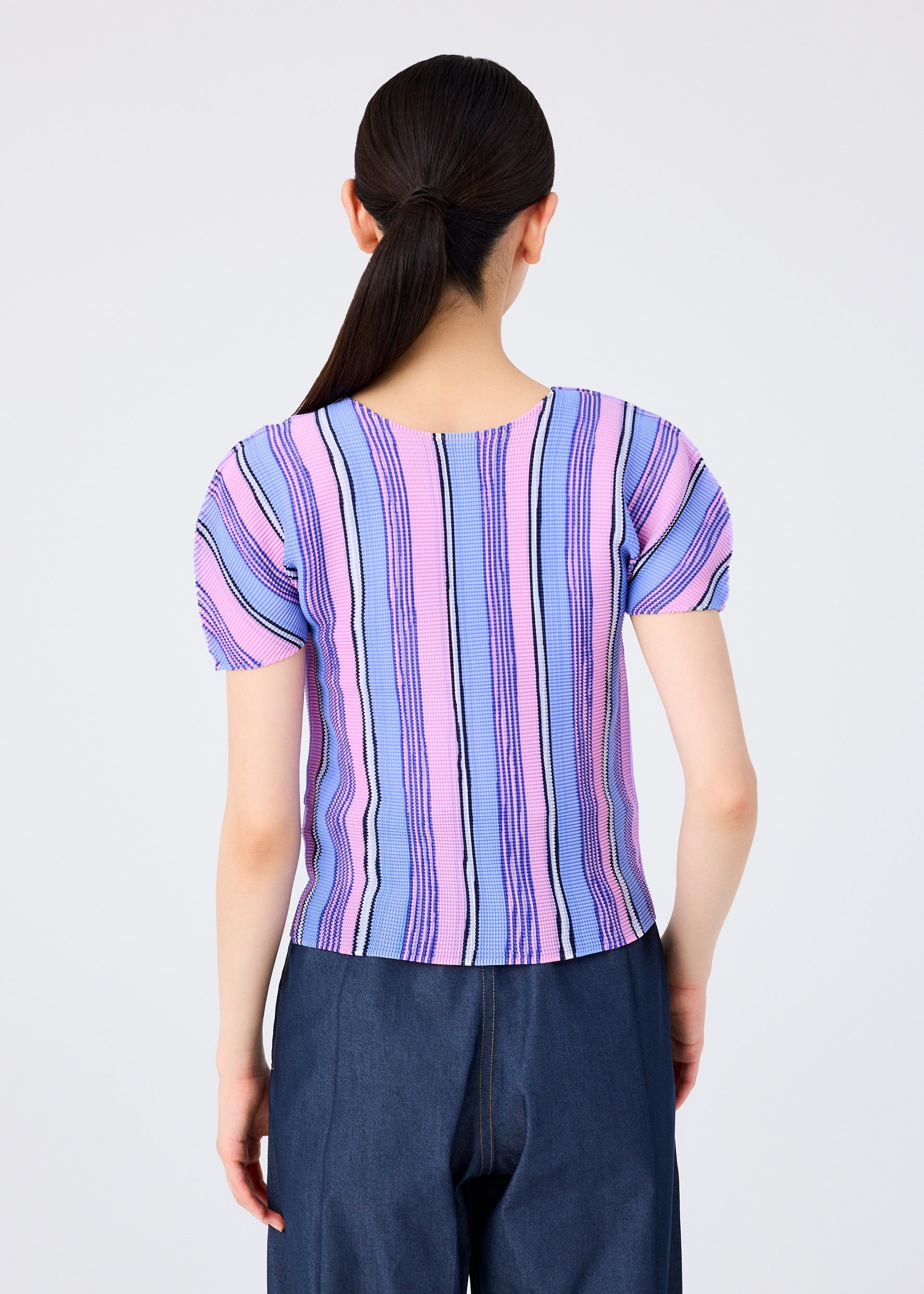 Zoomed view A person with long dark hair in a low ponytail wears the me ISSEY MIYAKE SHIRT STRIPE top in pastel pink, blue, and purple stripes with dark blue pants, standing against a plain white background.