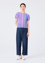 A woman with straight dark hair pulled back faces the camera in a me ISSEY MIYAKE SHIRT STRIPE short-sleeved top, featuring a pink and purple stripe pattern, paired with wide-leg dark blue pants and white slip-on shoes against a plain white background.