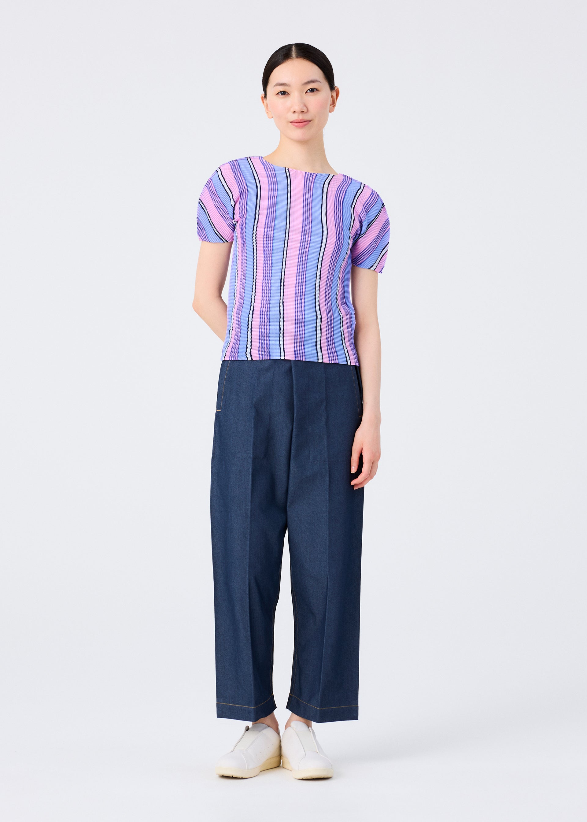 Zoomed view A woman with straight dark hair pulled back faces the camera in a me ISSEY MIYAKE SHIRT STRIPE short-sleeved top, featuring a pink and purple stripe pattern, paired with wide-leg dark blue pants and white slip-on shoes against a plain white background.