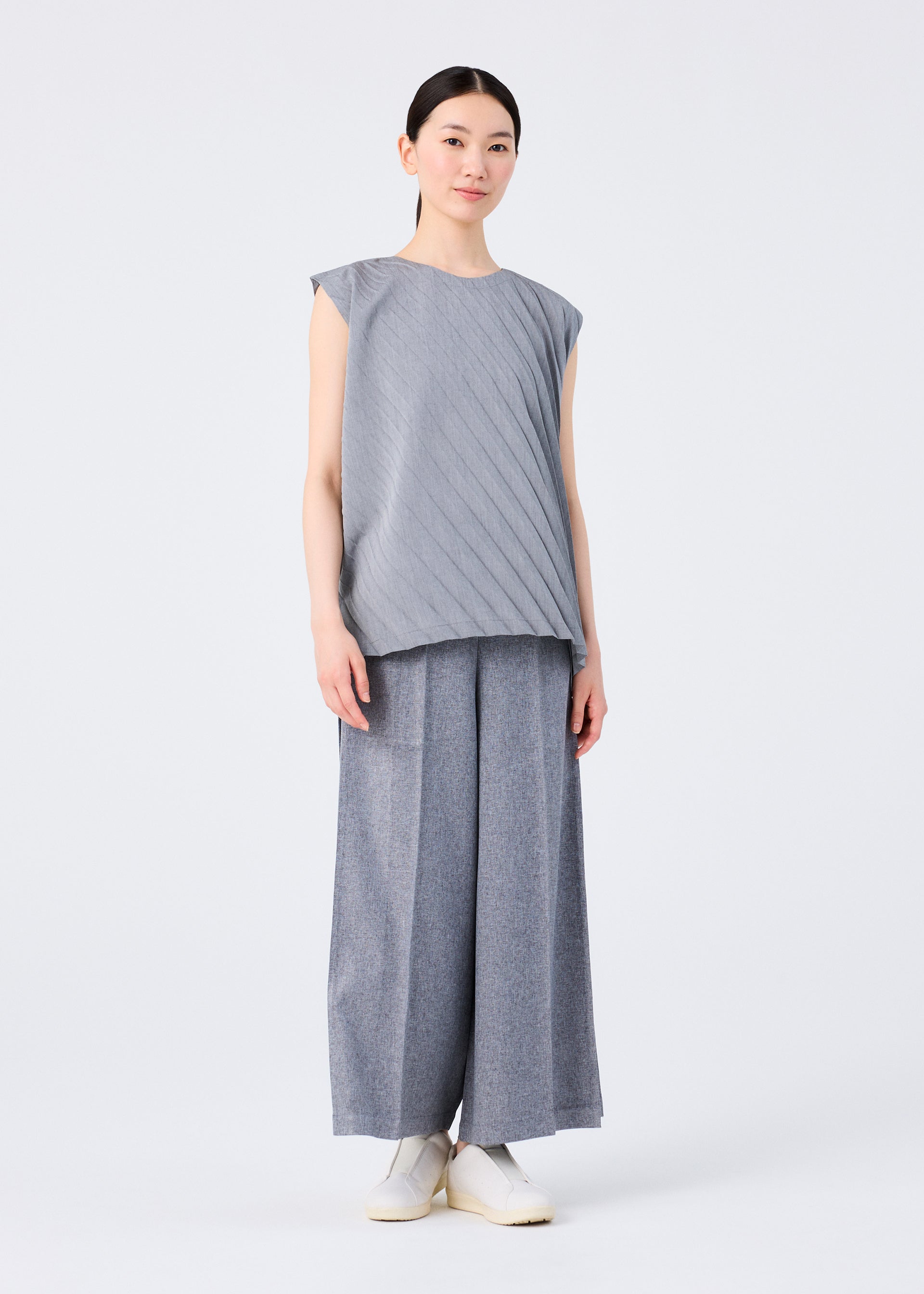 Zoomed view A woman with straight dark hair pulled back wears the me ISSEY MIYAKE ANGLE PLEATS SHIRT and wide-legged gray pants, styled with white slip-on shoes against a plain white background.