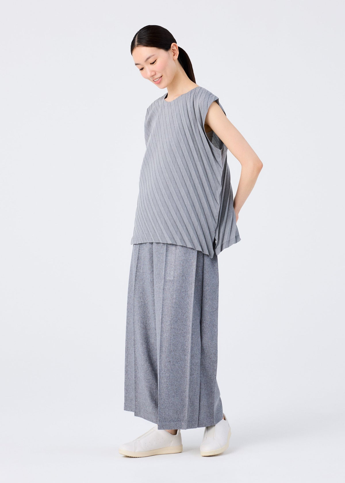 A woman stands against a white background, smiling slightly with her hands behind her back. She wears the loose-fitting ANGLE PLEATS SHIRT by me ISSEY MIYAKE, wide-legged gray pants, and white slip-on shoes. view 9 of 9