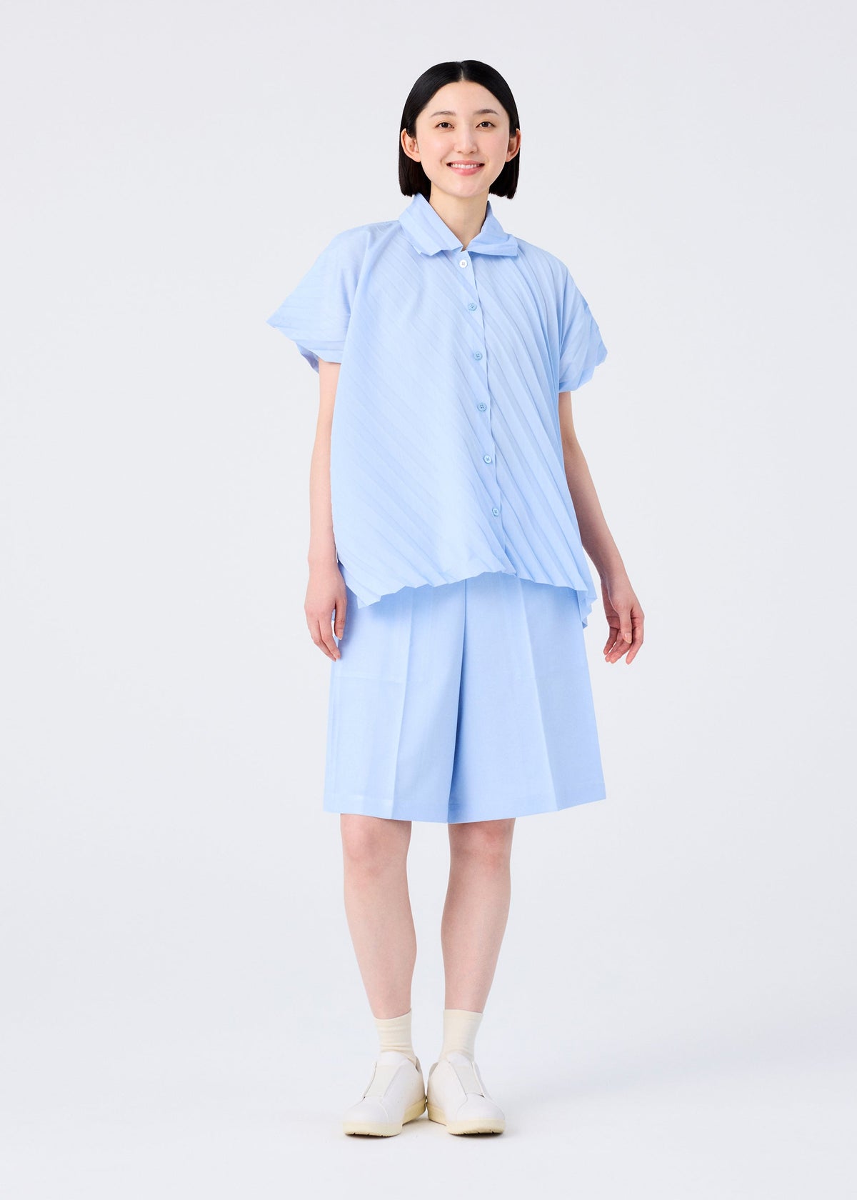 A smiling person with short black hair wears the me ISSEY MIYAKE ANGLE PLEATS SHIRT in light blue melange and matching knee-length shorts, with white socks and slip-on shoes, standing against a plain white background. view 8 of 9