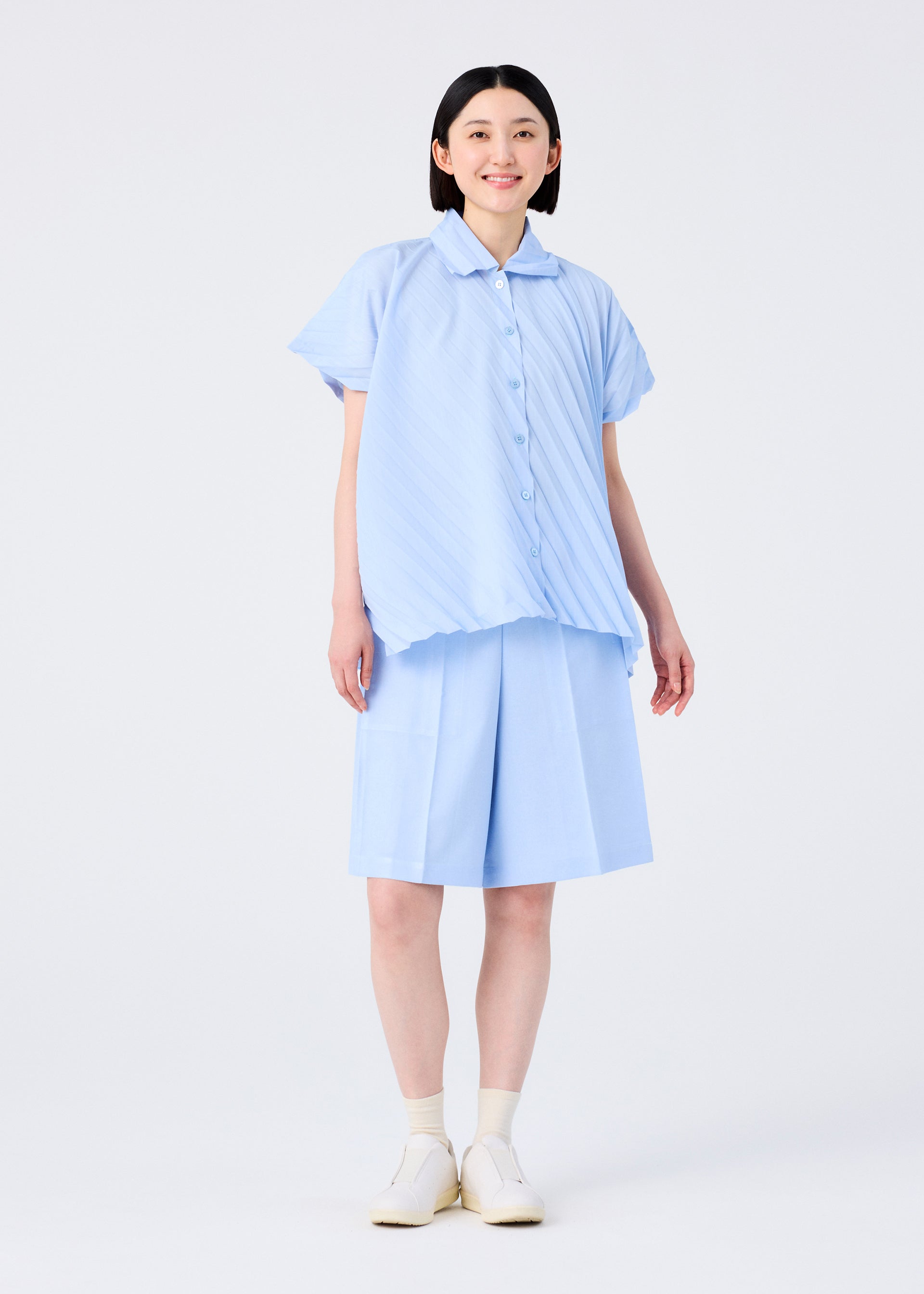 Zoomed view A smiling person with short black hair wears the me ISSEY MIYAKE ANGLE PLEATS SHIRT in light blue melange and matching knee-length shorts, with white socks and slip-on shoes, standing against a plain white background.