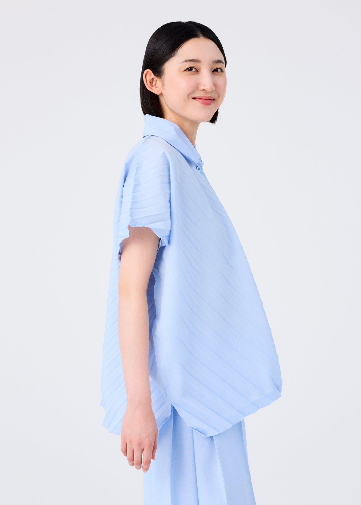 A person with straight dark hair smiles while wearing the me ISSEY MIYAKE ANGLE PLEATS SHIRT in a loose, light blue melange finish. They stand sideways against a plain white background. view 9 of 9