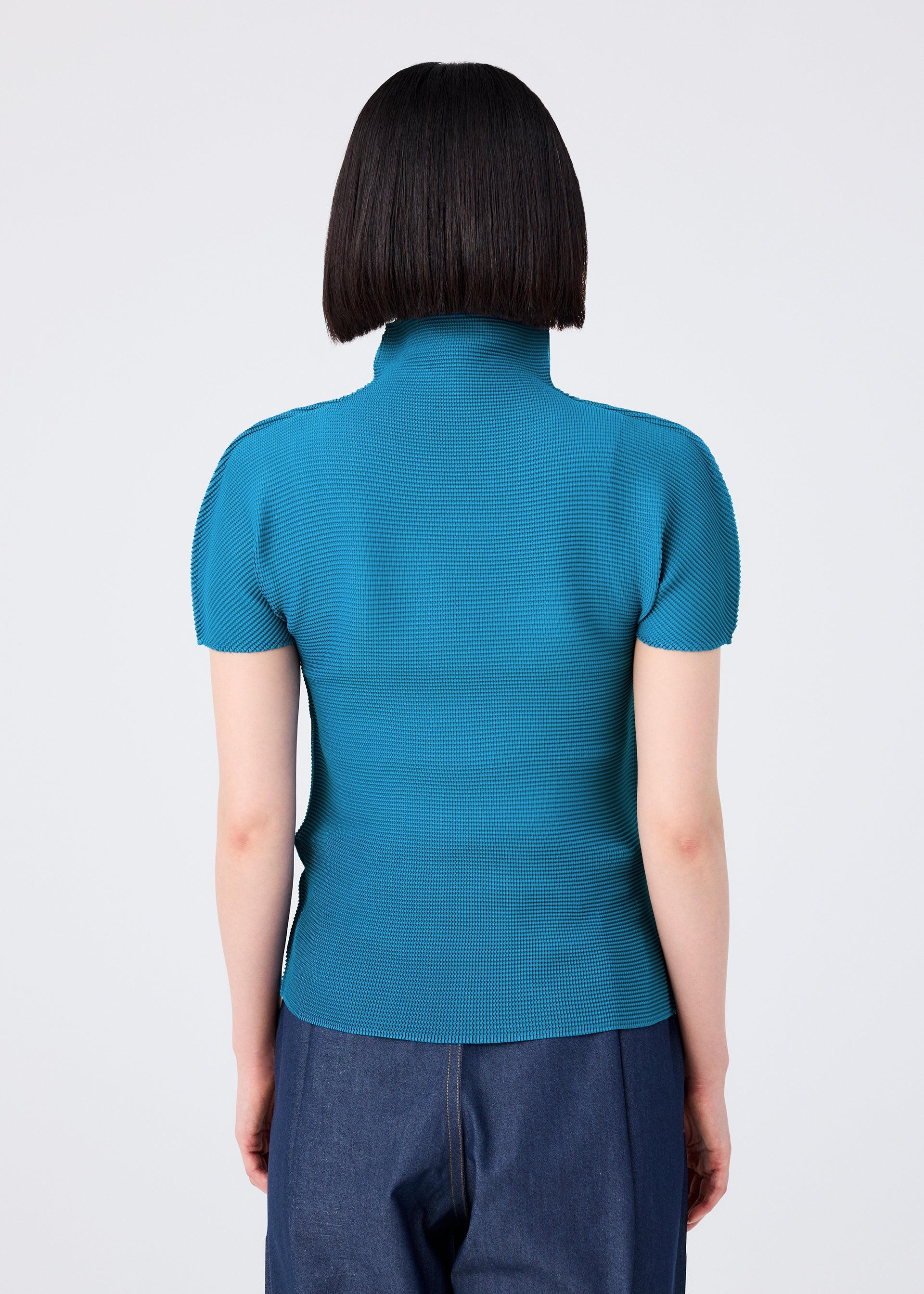 Zoomed view A person with straight, chin-length dark hair is seen from behind, wearing the STRETCH PLEATS 6 top by me ISSEY MIYAKE in teal with dark blue jeans, standing against a plain light background.