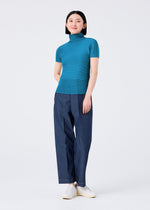 A woman smiles against a plain white background, wearing the me ISSEY MIYAKE STRETCH PLEATS 6 teal short-sleeved high-neck top, loose blue pants, and white shoes.