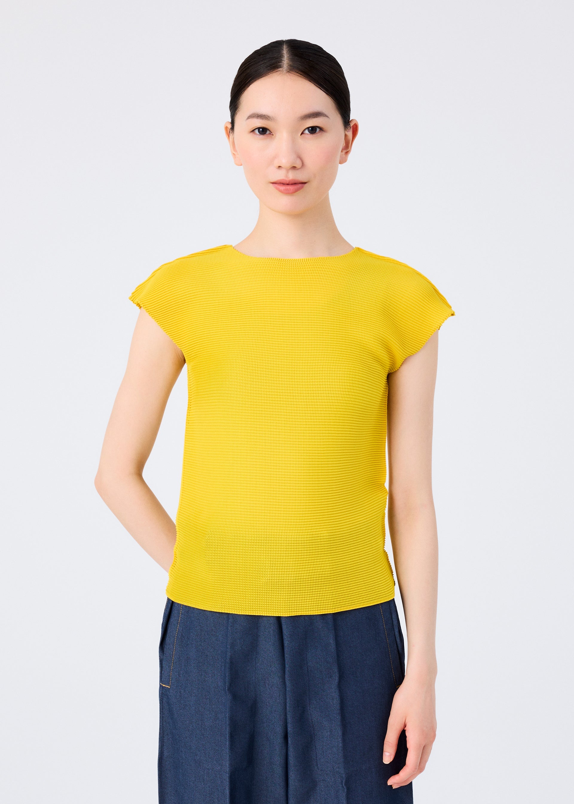 Zoomed view A woman with straight dark hair tied back wears a bright yellow STRETCH PLEATS 6 top from me ISSEY MIYAKE and high-waisted blue pants. She faces forward against a plain white background with a neutral expression.
