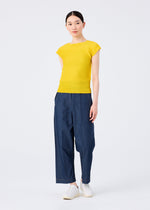 A woman models the me ISSEY MIYAKE STRETCH PLEATS 6: a fitted yellow round-neck top with French sleeves, paired with loose blue pants and white slip-on shoes. She stands against a white background, hair pulled back, looking slightly to the side.