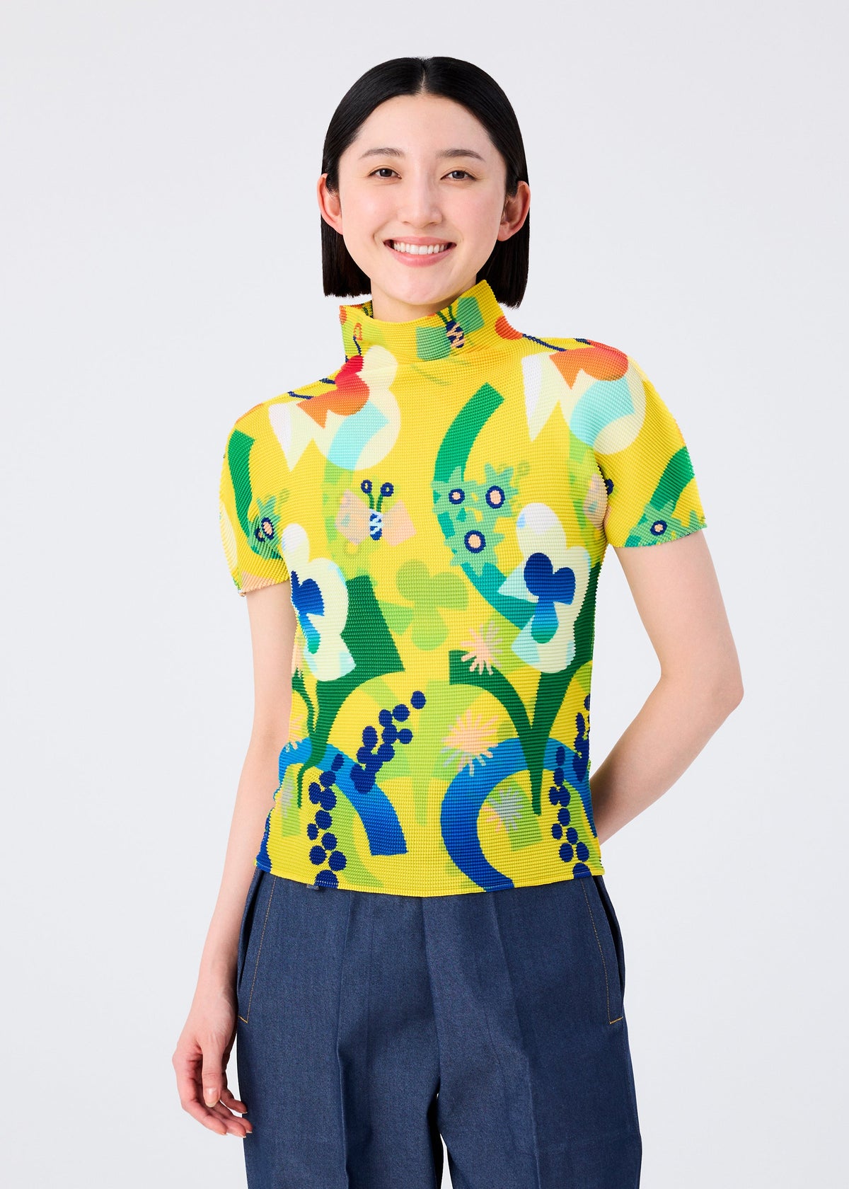 A woman with straight dark hair smiles while wearing the me ISSEY MIYAKE FLOWER DOME bright yellow high-neck top with a colorful floral pattern and high-waisted blue pants, standing against a plain white background. view 5 of 7