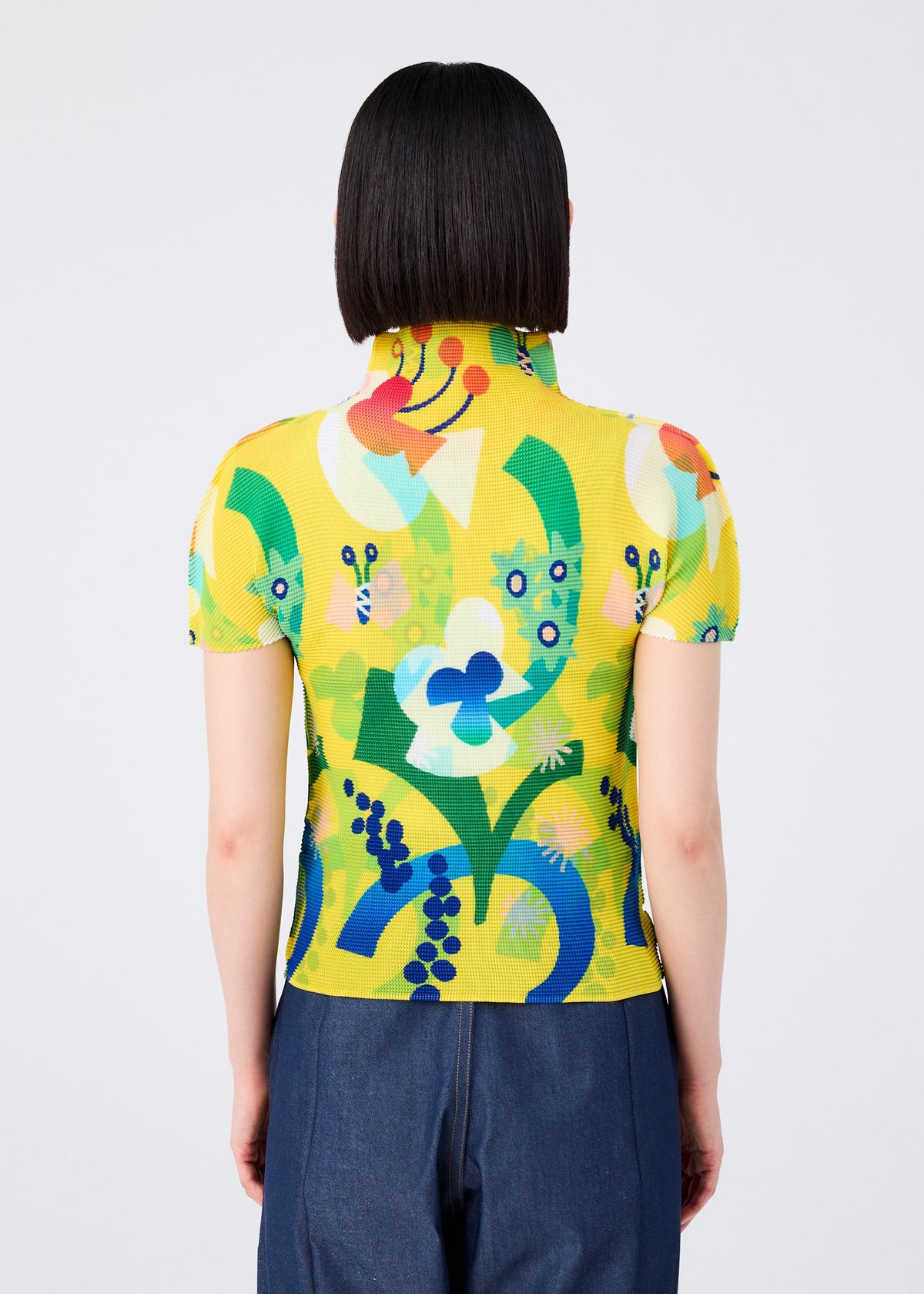 A person with straight, chin-length dark hair is seen from behind wearing the FLOWER DOME stretch pleats top by me ISSEY MIYAKE, featuring a colorful floral botanical pattern and blue pants against a plain light background. view 6 of 7