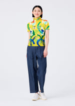 A person smiles in the FLOWER DOME yellow short-sleeve shirt by me ISSEY MIYAKE, featuring a bold floral botanical print, paired with loose blue pants and white slip-on shoes, posed against a plain white background.