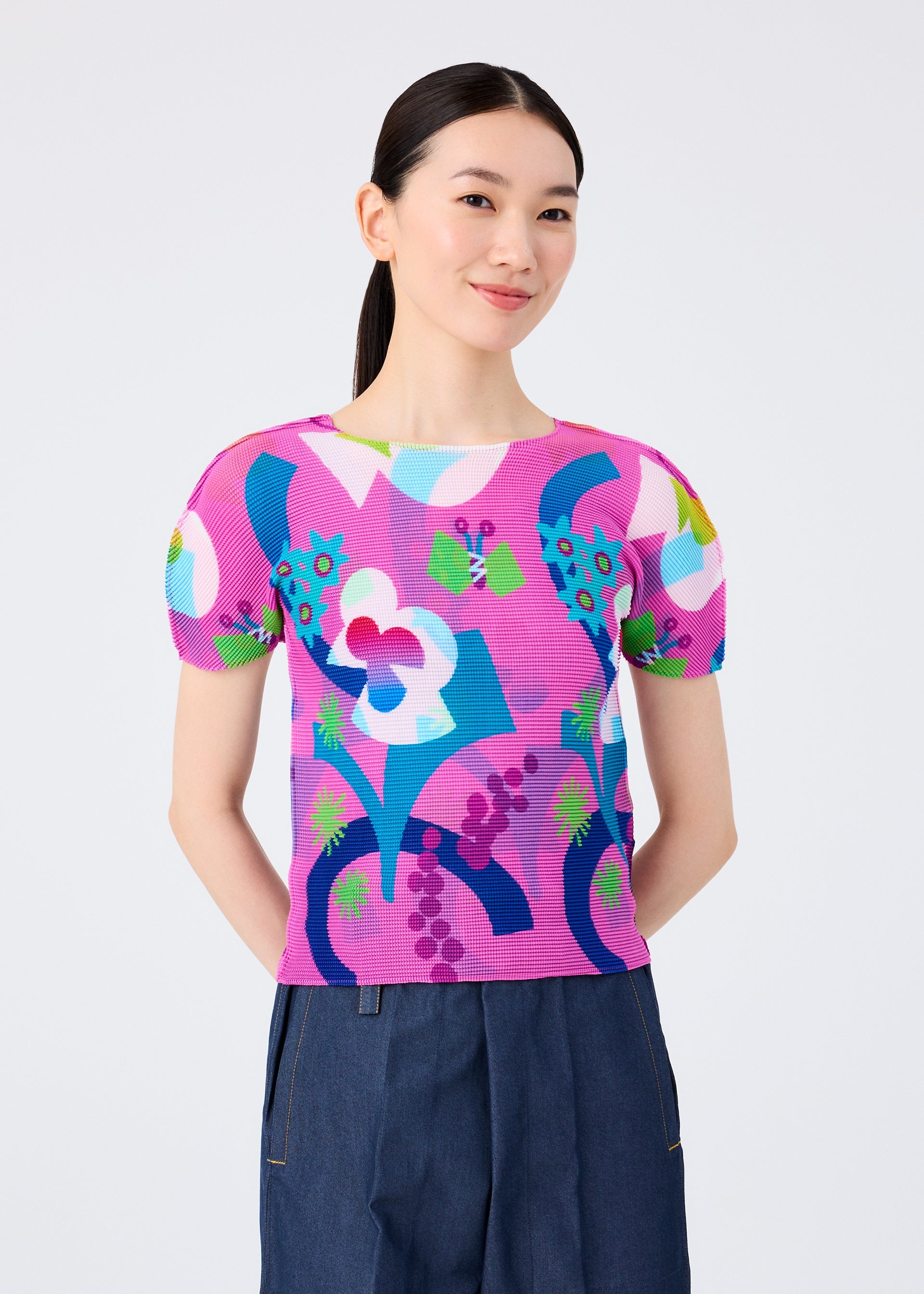 Zoomed view A woman with long dark hair in a ponytail smiles while wearing the FLOWER DOME top by me ISSEY MIYAKE and high-waisted blue pants, standing against a plain white background.