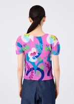 A person with long dark hair in a low ponytail is seen from behind wearing the me ISSEY MIYAKE FLOWER DOME top, a vibrant short-sleeve round-neck piece with bold pink, blue, green, and white botanical patterns, paired with dark blue pants.