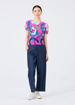 A person stands against a white background, smiling with hair tied back, wearing the me ISSEY MIYAKE FLOWER DOME top featuring bright floral and abstract patterns, paired with loose dark blue pants and white shoes.
