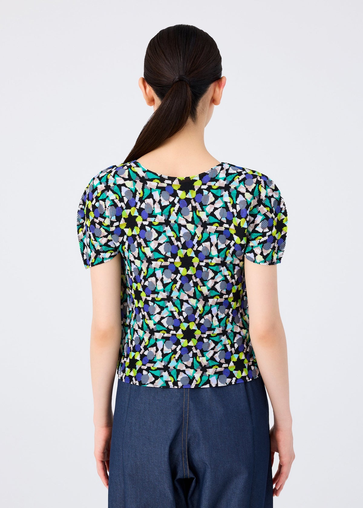 A person with long, dark hair in a low ponytail is seen from behind, wearing the me ISSEY MIYAKE KALEIDOSCOPE short-sleeved stretch pleats top and dark blue jeans against a plain light background. view 5 of 6