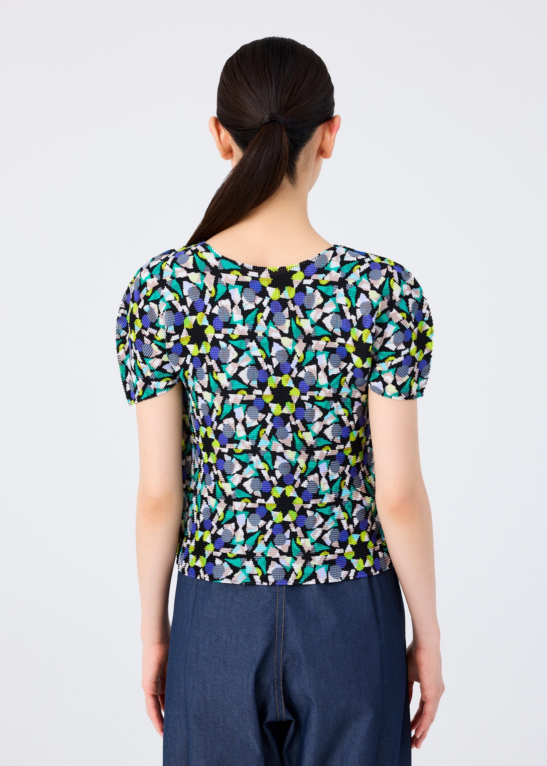 Zoomed view A person with long, dark hair in a low ponytail is seen from behind, wearing the me ISSEY MIYAKE KALEIDOSCOPE short-sleeved stretch pleats top and dark blue jeans against a plain light background.