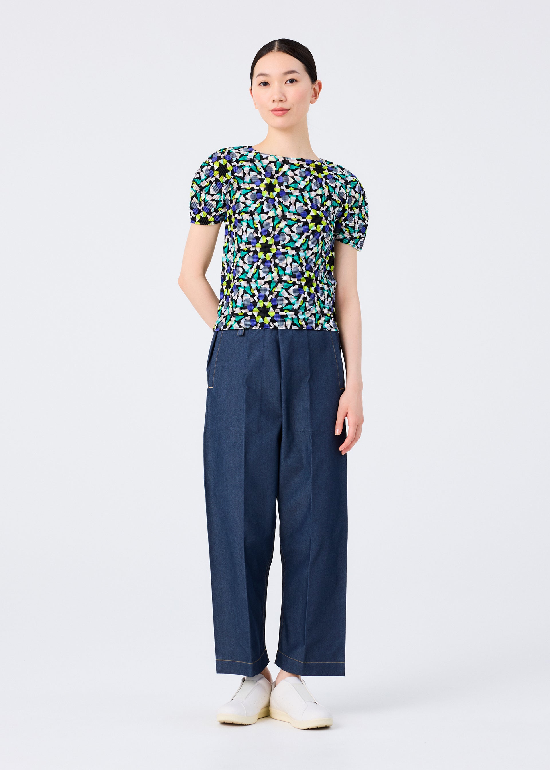 Zoomed view A woman faces forward against a white background, wearing the me ISSEY MIYAKE KALEIDOSCOPE short-sleeved stretch pleats top with green, blue, and yellow patterns, loose dark blue pants, and white slip-on shoes. Her arms are relaxed.
