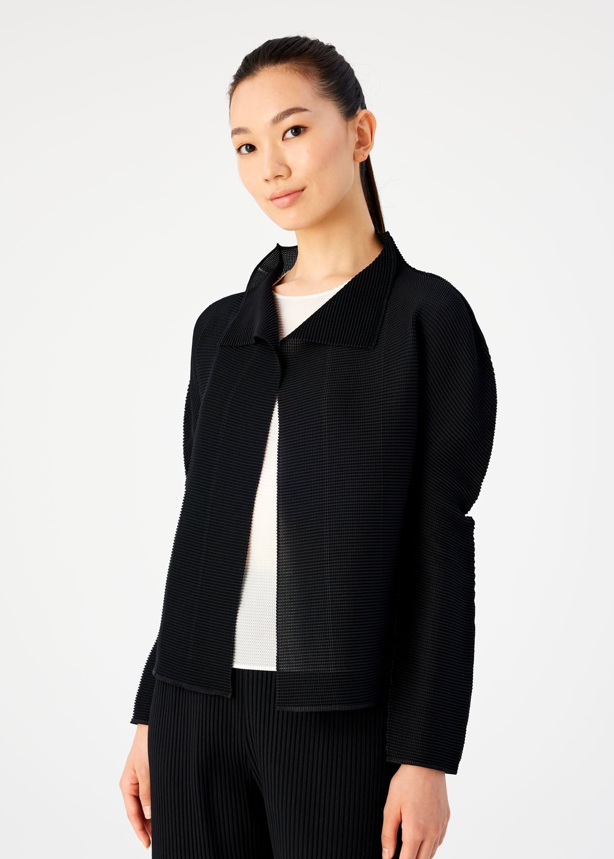 A woman with long dark hair tied back confidently models a black STRETCH PLEATS cardigan by me ISSEY MIYAKE over a white top and black pants, standing against a plain light background and looking at the camera. view 4 of 6