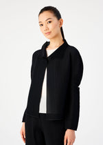 A woman with long dark hair tied back confidently models a black STRETCH PLEATS cardigan by me ISSEY MIYAKE over a white top and black pants, standing against a plain light background and looking at the camera.