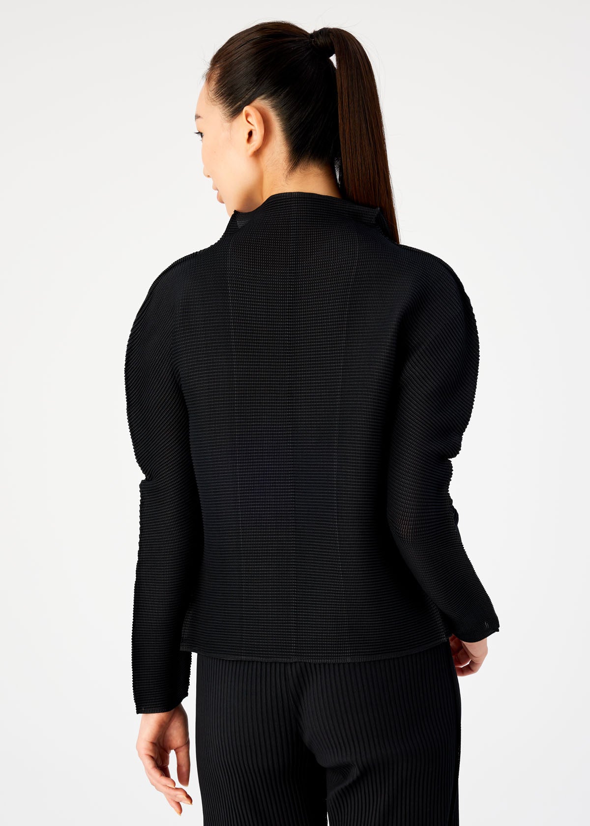 Zoomed view A woman with long dark hair in a ponytail wears the me ISSEY MIYAKE STRETCH PLEATS jacket and pants, standing with her back to the camera against a plain light background.