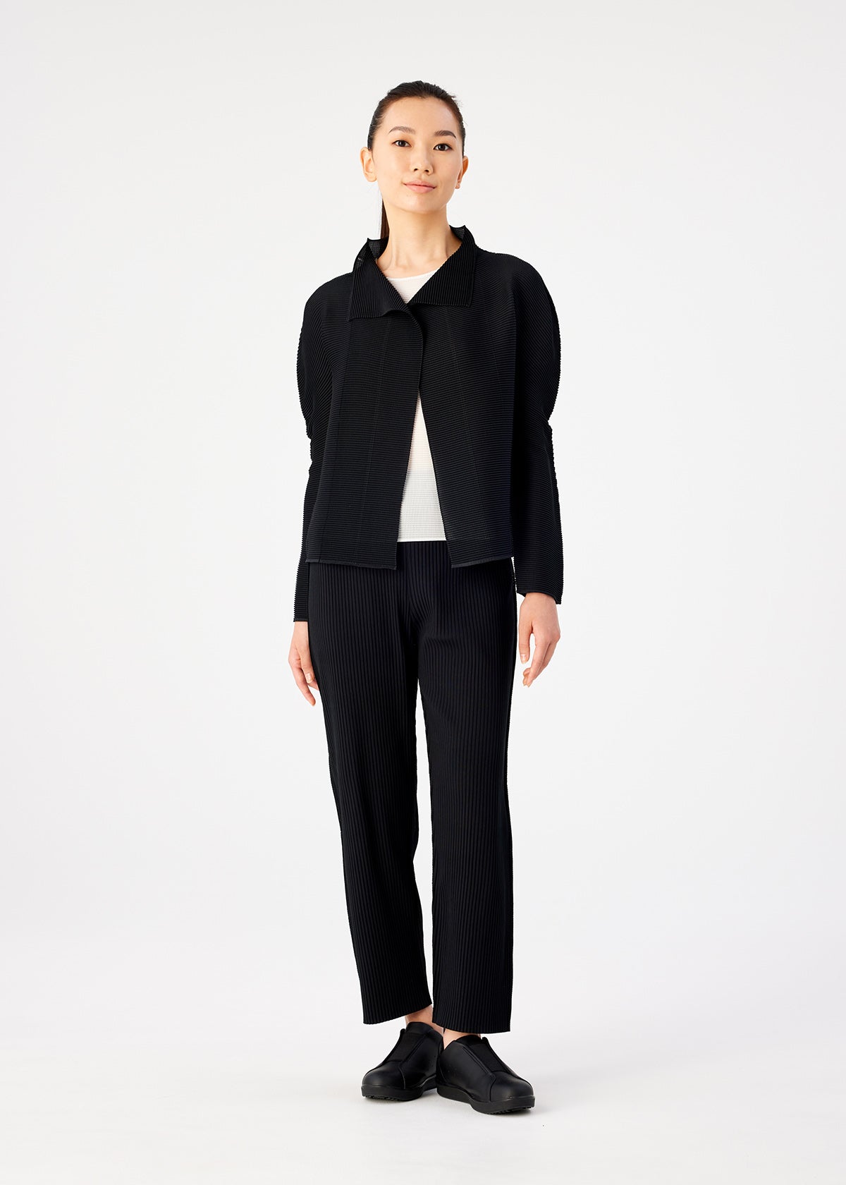A person stands facing forward against a white background, wearing me ISSEY MIYAKE STRETCH PLEATS black cardigan and pleated pants, a white top, and black shoes. Their hair is tied back and arms rest at their sides. view 6 of 6
