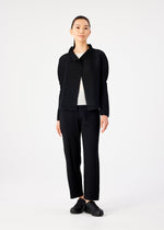 A person stands facing forward against a white background, wearing me ISSEY MIYAKE STRETCH PLEATS black cardigan and pleated pants, a white top, and black shoes. Their hair is tied back and arms rest at their sides.