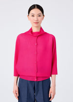 A woman faces forward in a bright pink me ISSEY MIYAKE DOLMAN STRETCH PLEATS 1 cardigan with a draped collar and loose fit, paired with dark blue pants. The background is plain white.